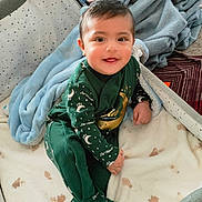 Danish is registered to the contest to win money with this photo: baby, child, infant, crib, blanket, pillow, bed, smile, happy, indoors, clothing, green_outfit, footed_pajamas, face, person, soft_texture, sleepwear, cute, toddler, cozy