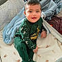 baby, child, infant, crib, blanket, pillow, bed, smile, happy, indoors, clothing, green_outfit, footed_pajamas, face, person, soft_texture, sleepwear, cute, toddler, cozy