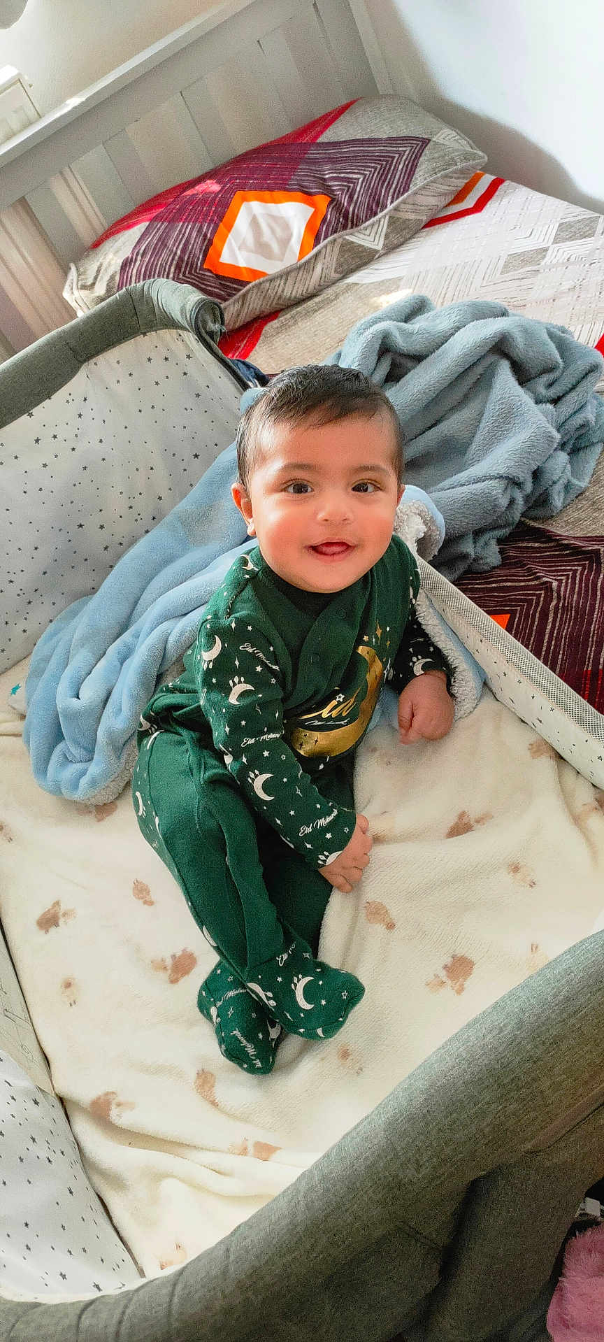 Danish is registered to the contest to win money with this photo: baby, child, infant, crib, blanket, pillow, bed, smile, happy, indoors, clothing, green_outfit, footed_pajamas, face, person, soft_texture, sleepwear, cute, toddler, cozy
