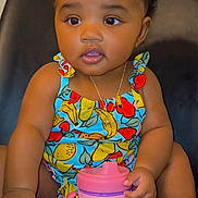 Marley is registered to the contest to win money with this photo: toddler, child, baby, sitting, colorful_clothing, hair_bows, sippy_cup, fruit_pattern, curious, indoors, portrait, cute, small_hands, necklace, black_hair, chair, bright_eyes, skin, face, expression