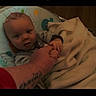 adult_hand, baby, black_border, blanket, candid, child, cozy, face, home, indoor, infant, looking_at_camera, open_mouth, patterned_seat, portrait, sitting, tattoo, text_overlay, warm_lighting, wide_eyed