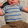 baby, child, toy, train, striped_shirt, brown_pants, bed, blanket, smiling, tongue_out, person, cute, infant, playful, indoors, happy, blue_eyes, short_hair, clothing, lying_down