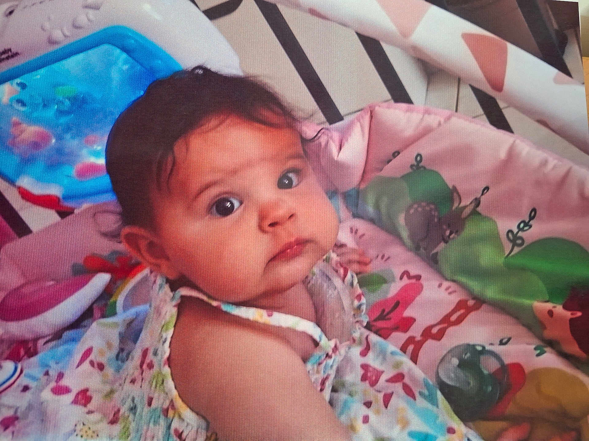élise participe au concours pour gagner de l'argent avec cette photo : baby, child, infant, playmat, toys, colorful, floral_dress, curious, indoors, cute, face, person, young_child, expression, lying_down, soft_toys, play_area, head, skin, eyes