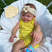 Amaria joined the competition — help win amazing prizes! baby, child, yellow_outfit, headband, flower, smile, sitting, blanket, outdoor, grass, wooden_sign, footwear, sandal, cute, infant, happy, person, skin, head, nature