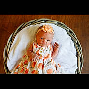 Georgia is registered to the contest to win money with this photo: baby, infant, flower_headband, dress, basket, blanket, wooden_floor, portrait, cute, child, newborn, orange_pattern, headband, soft_texture, indoor, lying_down, adorable, young_child, peaceful, cozy