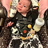 baby, infant, car_seat, pacifier, milk_bottle, black_clothing, white_clothing, cow_print, toy, wooden_beads, harness, cute, expression, pink_background, patterned_fabric, sitting, child, indoor, face, playful