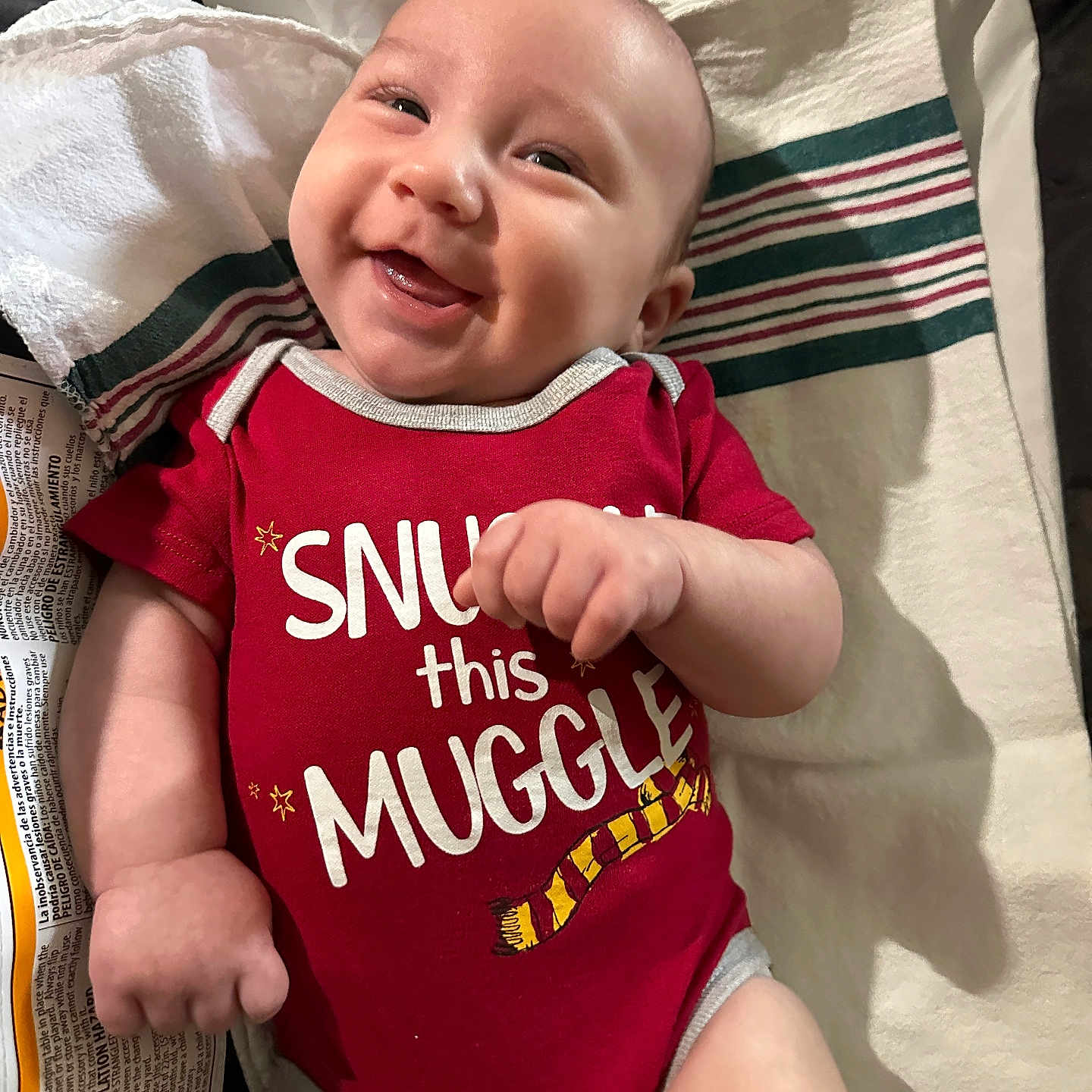 Luke joined the competition — help win amazing prizes! arm, baby, child, clothing, cute, expression, face, hand, happy, indoors, infant, lying_down, newborn, person, portrait, red_onesie, skin, smile, striped_cloth, text_on_clothing