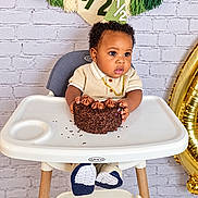 Davier is registered to the contest to win money with this photo: baby, child, birthday, cake, chocolate_cake, high_chair, smiling, portrait, indoor, celebration, curly_hair, gold_chain, cream_outfit, blue_shoes, sprinkles, frosting, balloon, brick_wall, party_decorations, photography