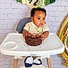 baby, child, birthday, cake, chocolate_cake, high_chair, smiling, portrait, indoor, celebration, curly_hair, gold_chain, cream_outfit, blue_shoes, sprinkles, frosting, balloon, brick_wall, party_decorations, photography