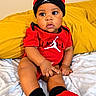 Davier is registered to the contest to win money with this photo: baby, child, infant, portrait, sitting, bed, blanket, pillow, red_clothing, black_hat, beanie, jordan_style, sportswear, socks, shoes, indoors, bedroom, yellow_background, looking_at_camera, close_up
