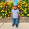 child, toddler, portrait, outdoor, garden, flowers, marigolds, dinosaur_hat, blue_hat, smiling, standing, facial_expression, gray_shirt, jeans, sneakers, sidewalk, stone_wall, plants, daylight, photography