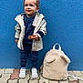 toddler, child, cookie, blue_wall, backpack, white_sneakers, beige_jacket, navy_clothing, sidewalk, outdoor, cute, smiling, standing, texture, pavement, fashion, accessory, playful, small_person, casual