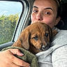 puppy, dog, person, woman, face, portrait, cuddling, car, vehicle, passenger_seat, towel, blanket, hand, ring, sunglasses, window, outdoor, trees, cute, sleepy