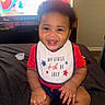 baby, smiling, bib, red_shirt, sitting, indoor, television, blurred_background, child, happy, face, hands, shorts, home, cute, infant, celebration, holiday, portrait, candid