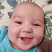 Layne is registered to the contest to win money with this photo: baby, bed, blue_shirt, cheeks, child, close_up, cute, eyes, face, floral_bedding, happy, indoors, infant, lying_down, open_mouth, plush_toy, portrait, smiling, soft_lighting, stuffed_animal