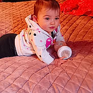 Supreme is registered to the contest to win money with this photo: baby, bottle, bed, blanket, clothing, curly_hair, face, furniture, hair_tie, indoors, infant, person, pink, quilted_bedspread, sleeves, soft_lighting, toddler, toy, warm_colors, young_child