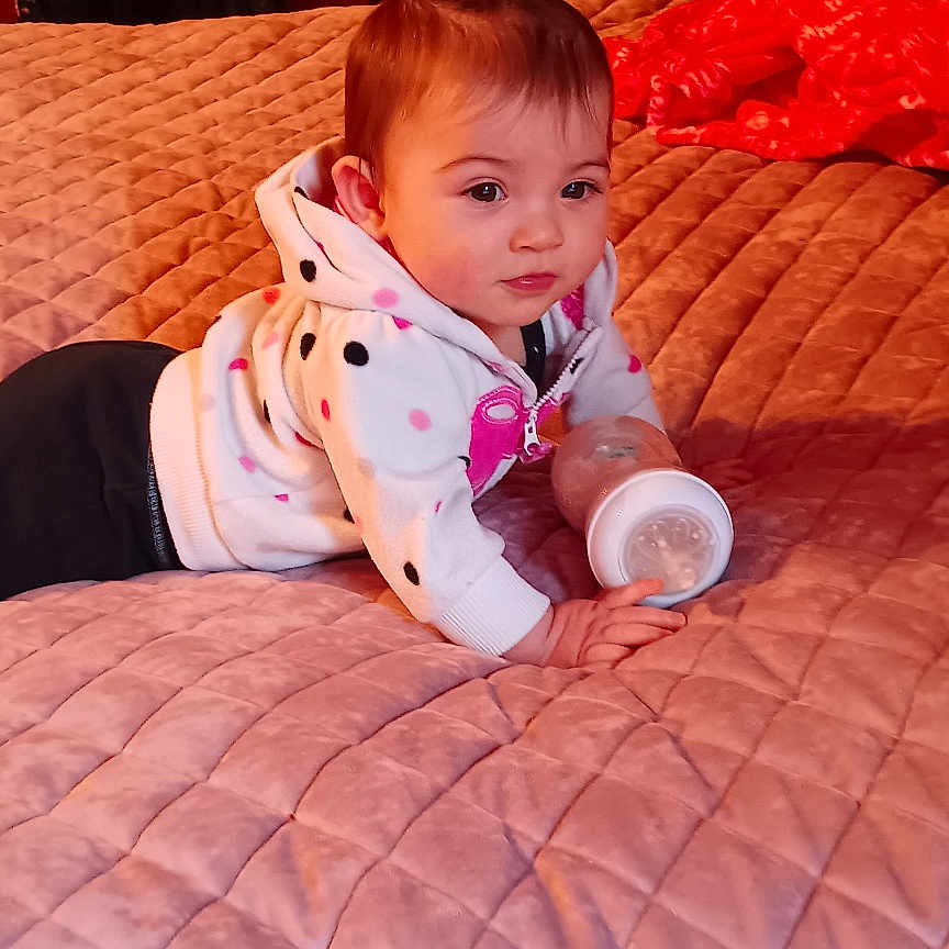 Supreme is registered to the contest to win money with this photo: baby, bottle, bed, blanket, clothing, curly_hair, face, furniture, hair_tie, indoors, infant, person, pink, quilted_bedspread, sleeves, soft_lighting, toddler, toy, warm_colors, young_child