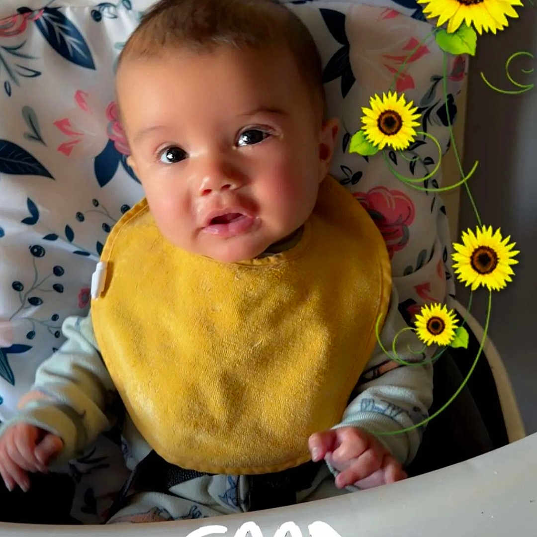 Mark joined the competition — help win amazing prizes! baby, child, high_chair, yellow_bib, face, expression, sunflowers, floral_pattern, cushion, indoor, person, smiling, morning, text_overlay, cute, infant, clothing, happy, portrait, feeding_time