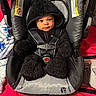baby, child, car_seat, black_clothing, hooded_jacket, straps, blanket, red_background, cute, infant, face, seat_belt, indoor, soft_texture, warm_clothing, looking_at_camera, portrait, cozy, young_child, smiling