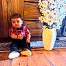 toddler, child, indoor, wood_floor, wooden_furniture, red_shirt, bow_tie, black_pants, white_shoes, vase, white_flowers, curious_expression, sitting, portrait, decor, home_interior, floor_rug, plush, cute, young_child