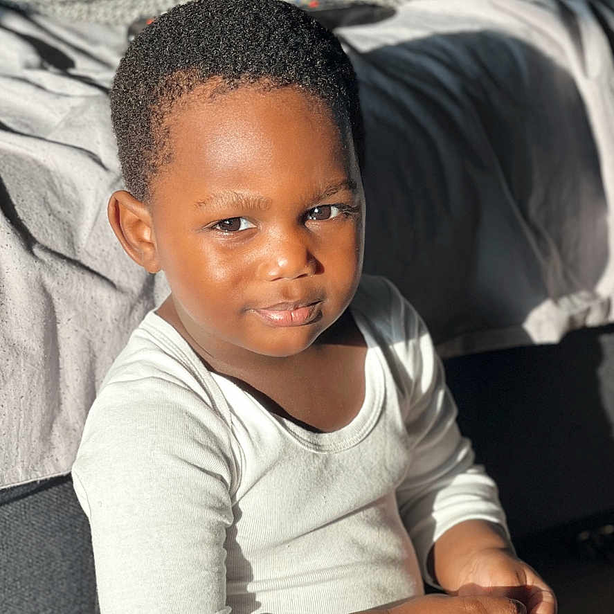 Nael participe au concours pour gagner de l'argent avec cette photo : bed, blanket, boy, casual, child, clothing, cute, face, floor, happy, indoor, long_sleeve, pants, person, portrait, relax, sitting, smile, sunlight, young