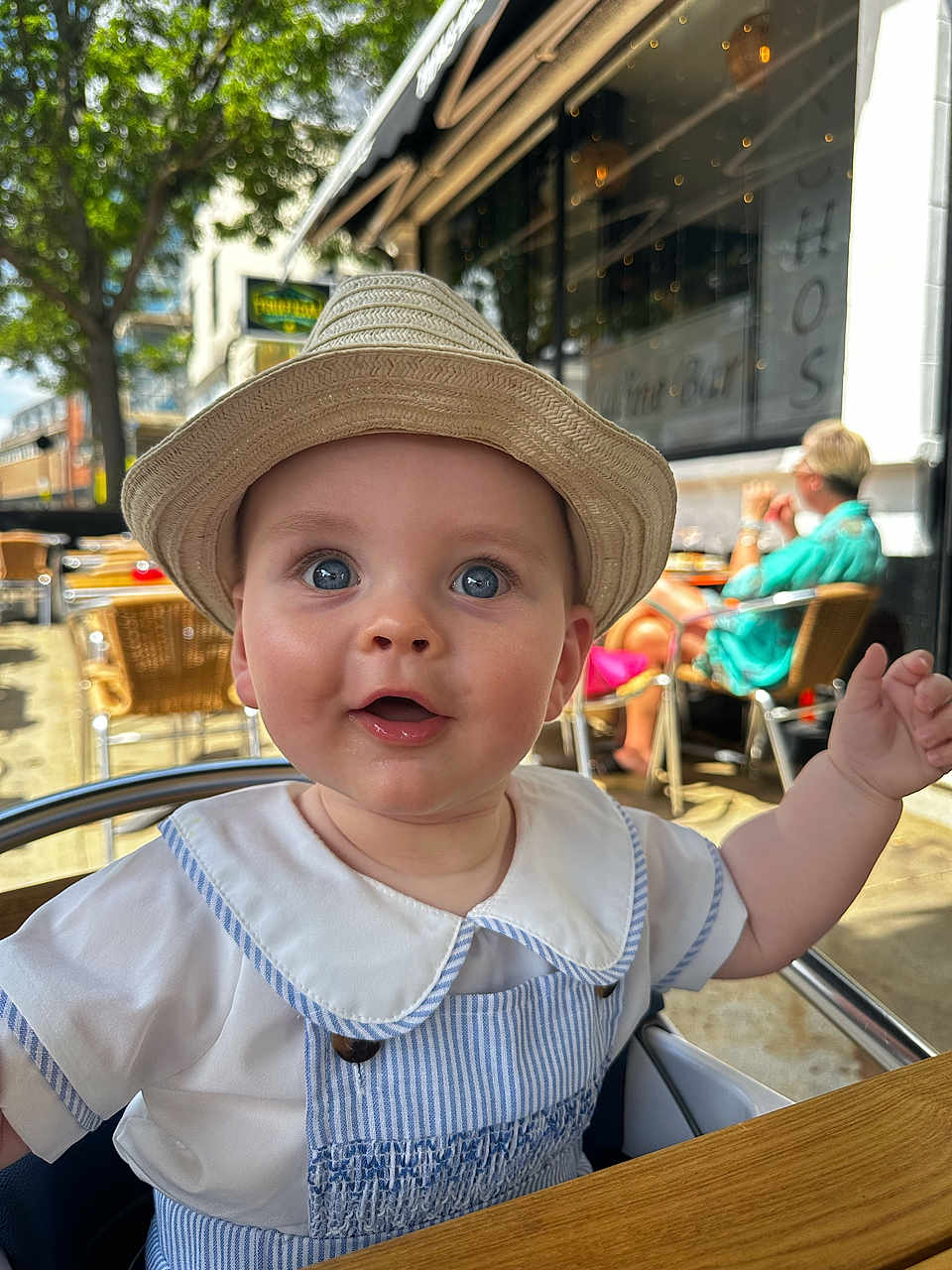 Woody is registered to the contest to win money with this photo: baby, child, hat, blue_eyes, outdoor, cafe, table, smile, person, clothing, seat, daylight, happy, face, young_child, summer, cute, people, background, portrait