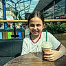 child, girl, smile, drink, starbucks, beverage, table, indoor, mall, shopping_center, glass_ceiling, blue_beams, plant, wooden_table, white_shirt, red_trim, happy, portrait, casual, daylight