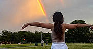 Natalia is registered to the contest to win money with this photo: child, rainbow, double_rainbow, grass, park, sky, clouds, evening, nature, outdoors, tree, person, standing, arms_outstretched, shorts, tank_top, casual, footwear, hair, scenery