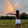 child, rainbow, double_rainbow, grass, park, sky, clouds, evening, nature, outdoors, tree, person, standing, arms_outstretched, shorts, tank_top, casual, footwear, hair, scenery