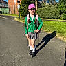 child, girl, smiling, backpack, pink_cap, green_cardigan, gray_skirt, white_socks, black_shoes, braided_hair, sidewalk, shadow, brick_building, greenery, bushes, tree, street_sign, sunny, outdoor, daytime