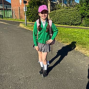Natalia is registered to the contest to win money with this photo: child, girl, school_uniform, pink_cap, backpack, braids, street, sidewalk, brick_building, greenery, sunlight, shadow, white_socks, black_shoes, grass, tree, outdoor, daylight, casual, smiling