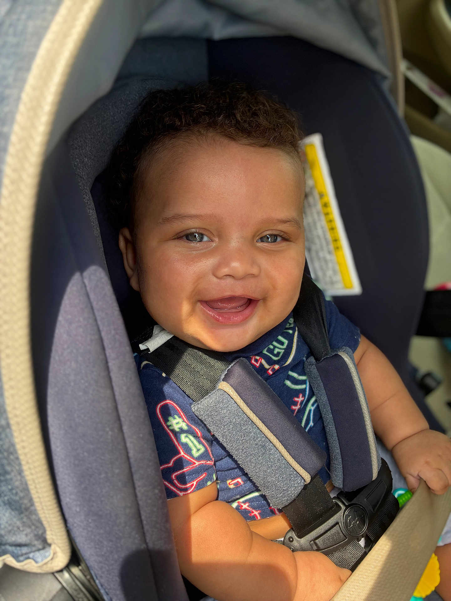 Amari is registered to the contest to win money with this photo: baby, blue_shirt, car_seat, child, close_up, curly_hair, cute, expression, hand, happy, indoor, infant, person, portrait, safety_harness, seat, seat_belt, skin, smile, sunlight