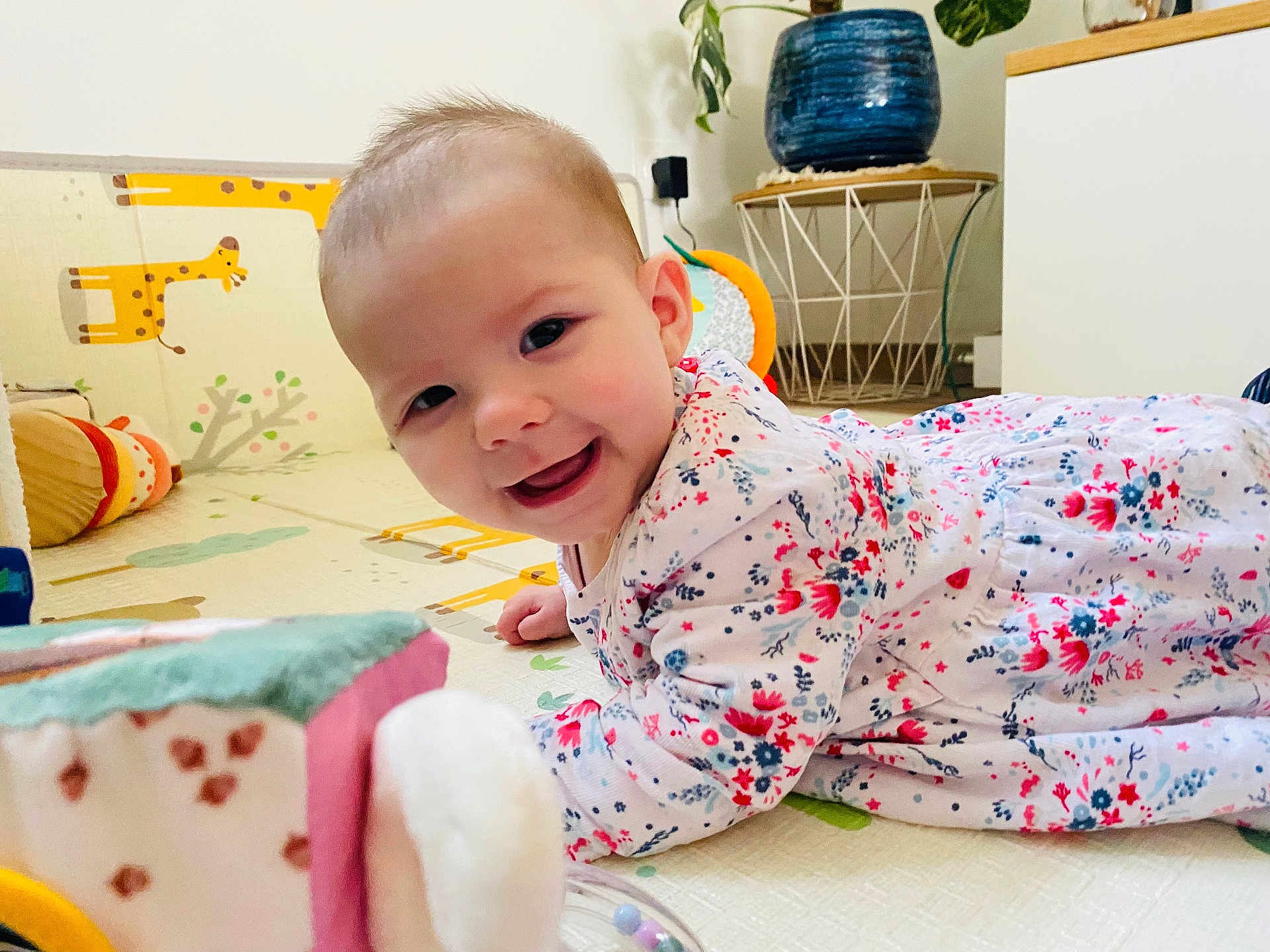 Charlie participe au concours pour gagner de l'argent avec cette photo : baby, child, smiling, play_mat, floral_clothing, indoor, toy, giraffe_illustration, face, happy, crawling, person, head, home_decor, plant, pot, floor, cute, infant, bright