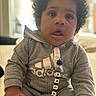 Malakai is registered to the contest to win money with this photo: adidas, baby, casual, child, clothing, curly_hair, cute, expression, face, grey_hoodie, hand, home, indoors, infant, pacifier_clip, person, portrait, sitting, soft_light, young_child