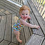 baby, child, blue_eyes, red_hair, onesie, wooden_deck, railing, standing, smiling, outdoor, sunlight, infant, happy, person, cute, summer, playful, barefoot, wood, colorful_clothing