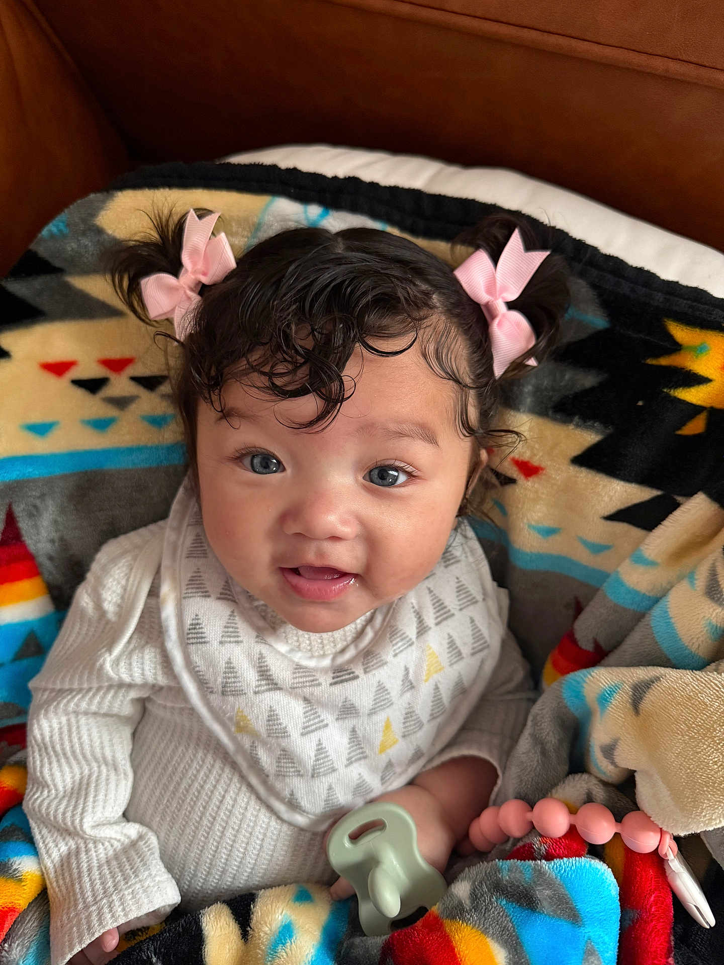 Avoni is registered to the contest to win money with this photo: baby, child, infant, bib, blanket, pink_bow, pigtails, pacifier, beaded_clip, couch, cozy, portrait, smile, drool, cheeks, curly_hair, onesie, hand, indoor, colorful_pattern