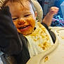 baby, high_chair, messy, food, bib, smile, happy, child, indoor, face, teeth, arm, clothing, eating, meal, plate, table, toy, background, person