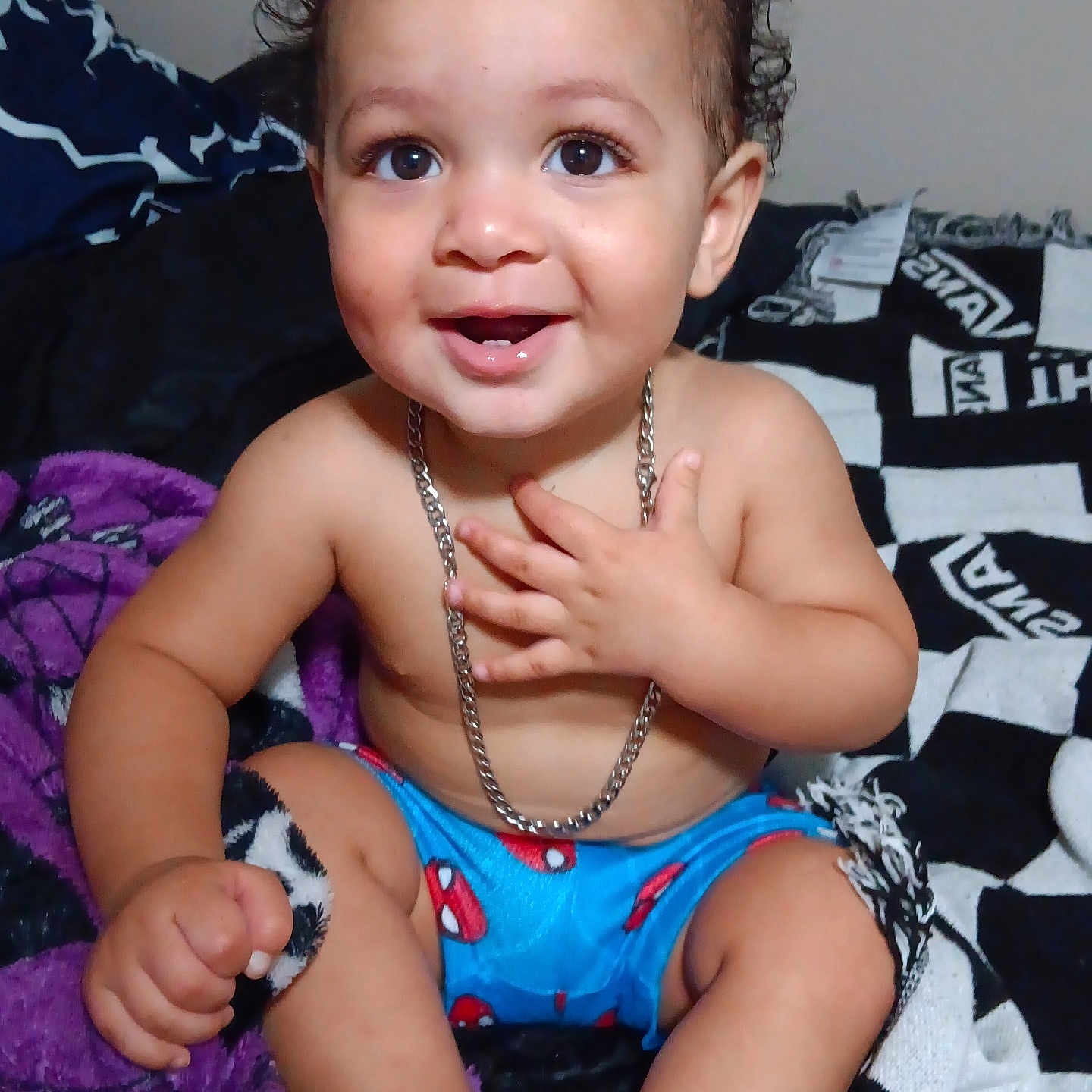 Zaylen is registered to the contest to win money with this photo: baby, child, toddler, smiling, happy, portrait, indoor, sitting, blanket, bed, necklace, curly_hair, brown_eyes, blue_shorts, bare_chested, hands, feet, close_up, infant, photography