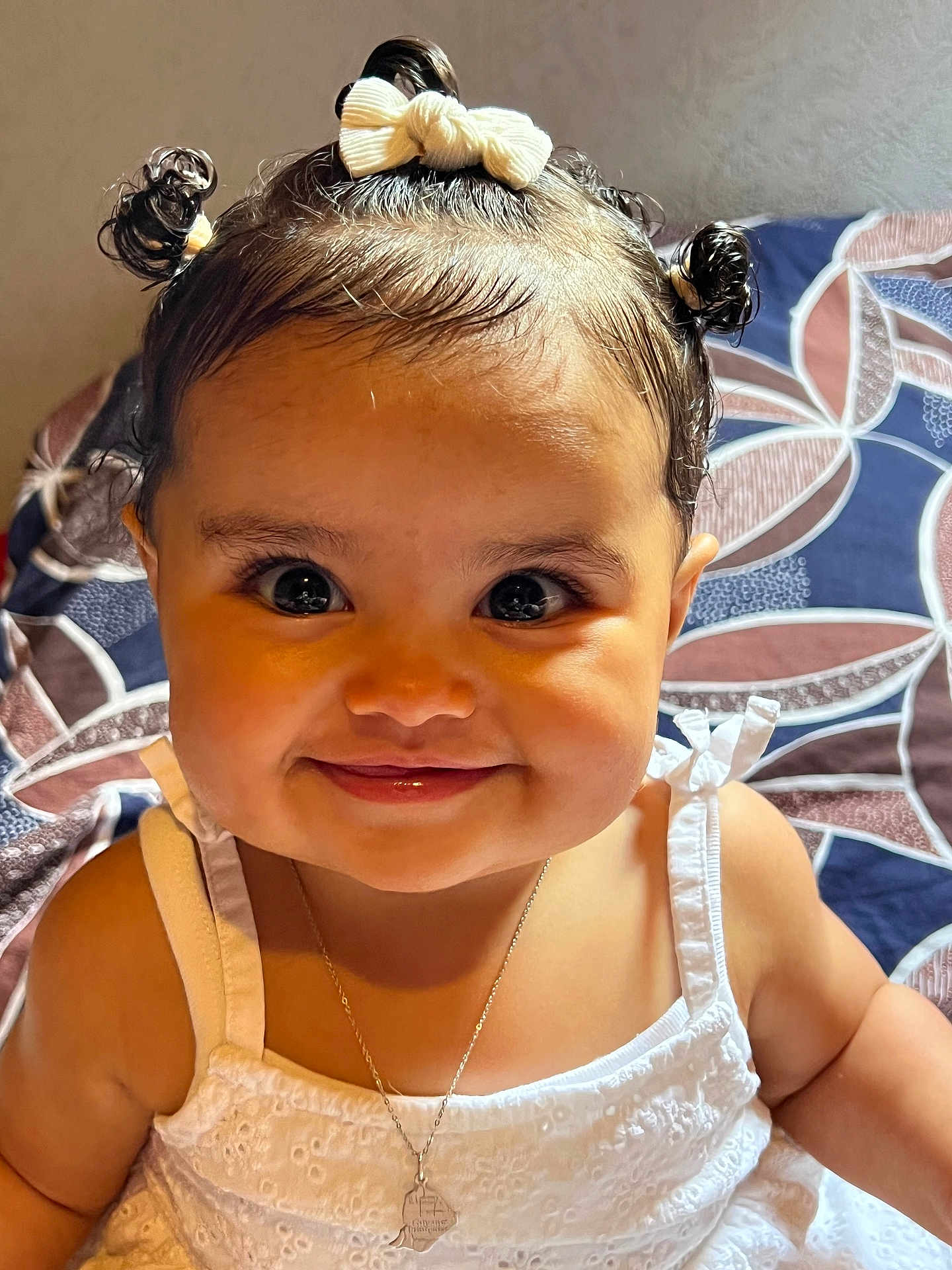 Amara a rejoint le concours — aidez-le/la à gagner de superbes lots ! baby, child, smile, face, dress, white_dress, hair_buns, bow, necklace, cute, indoors, cushion, patterned_background, portrait, happy, closeup, person, infant, young_child, skin