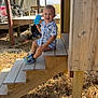 child, toddler, smile, outdoor, wooden_stairs, sippy_cup, shoes, sunlight, deck, grass, wood, person, playful, casual, daylight, happy, clothing, nature, summer, background