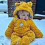 baby, infant, child, yellow_suit, hood, fur, snow, winter, sitting, blue_eyes, rosy_cheeks, portrait, outdoors, cold_weather, cute, brick_wall, blurred_background, plush, coat, boots