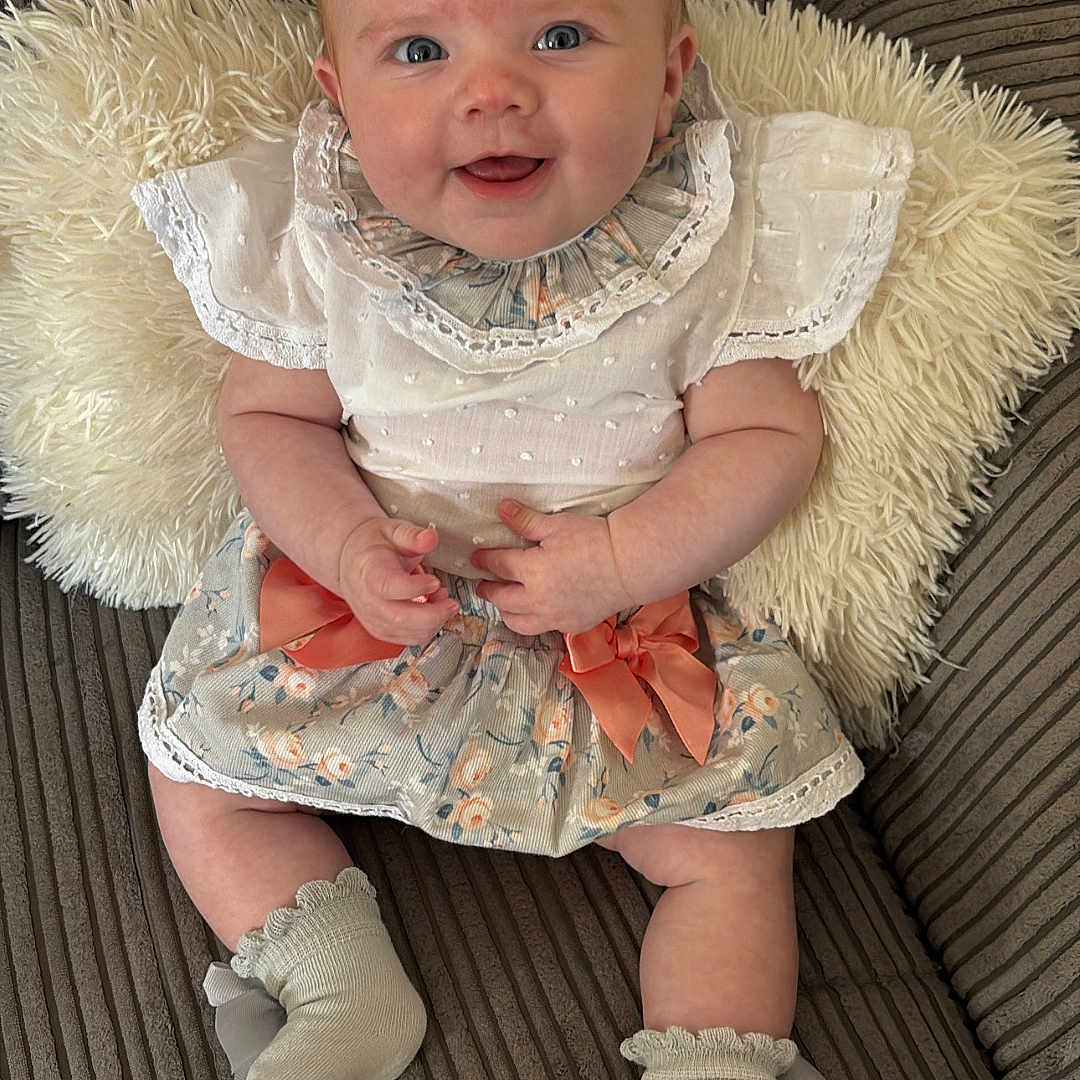 Myla is registered to the contest to win money with this photo: baby, bow, child, clothing, couch, cozy, cute, floral_skirt, happy, indoor, infant, person, pillow, portrait, seated, smiling, socks, soft_texture, striped_couch, white_top