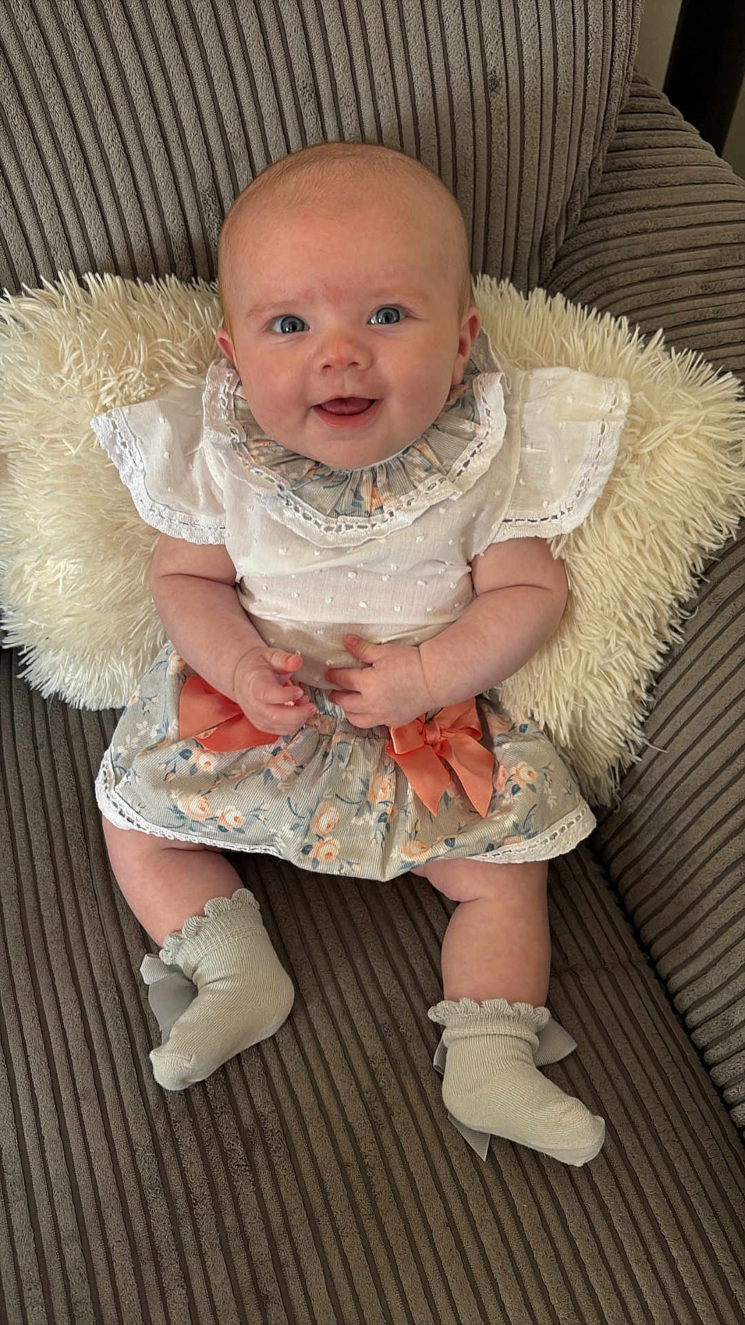 Myla is registered to the contest to win money with this photo: baby, smiling, couch, pillow, clothing, floral_skirt, bow, white_top, socks, cute, infant, happy, portrait, seated, indoor, child, person, soft_texture, striped_couch, cozy