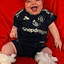 baby, infant, smiling, laughing, red_bow, headband, manchester_united, sportswear, black_shirt, white_socks, ruffled_socks, red_blanket, happy, cute, child, portrait, indoor, clothing, team_logo, fan