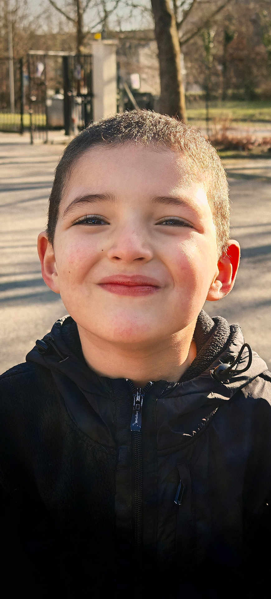 Haroun participe au concours pour gagner de l'argent avec cette photo : child, boy, smile, face, portrait, outdoor, jacket, black_clothing, short_hair, happy, sunlight, park, trees, fence, daytime, casual, young, person, head, warm