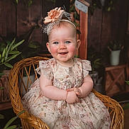 Ellie is registered to the contest to win money with this photo: baby, child, smiling, wicker_chair, floral_dress, headband, flower, rosy_cheeks, blue_eyes, indoor, plants, wooden_background, cute, portrait, sitting, happy, infant, decor, cozy, adorable
