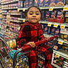 child, shopping_cart, grocery_store, pajamas, red, black, checkered_pattern, food_shelves, pasta_boxes, snacks, aisle, holding, sitting, young_child, casual_clothing, indoor, retail, supermarket, portrait, cute