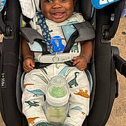 Khalil is registered to the contest to win money with this photo: baby, car_seat, smiling, dinosaur_print, pacifier, bottle, clothing, footwear, infant, child, seatbelt, happy, cute, indoor, person, baby_bottle, footwear_slippers, close_up, safety_harness, dinosaur_slippers