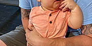Ace joined the competition — help win amazing prizes! baby, child, sunglasses, peach_shirt, shorts, tattoo, adult, hand, legs, casual, indoor, wood_floor, curious, fashion, person, seated, face, skin, expression, colorful