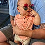 baby, child, sunglasses, peach_shirt, shorts, tattoo, adult, hand, legs, casual, indoor, wood_floor, curious, fashion, person, seated, face, skin, expression, colorful